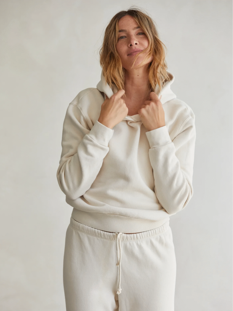 Woman in cozy off-white hoodie and sweatpants set, holding hood with relaxed expression.