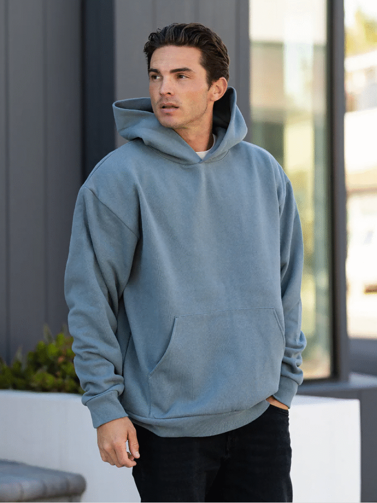 Man wearing oversized blue hoodie with front pocket, standing outdoors in casual urban setting.