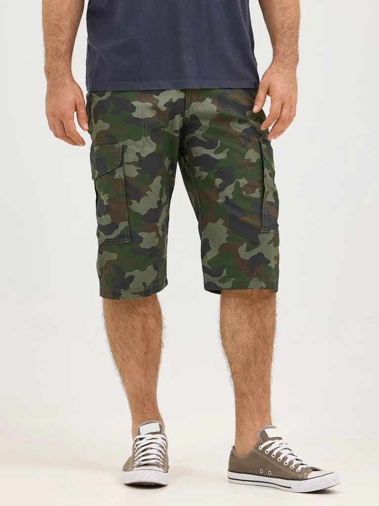 Man wearing knee-length camouflage cargo shorts with multiple side pockets, paired with a navy T-shirt and olive green low-top sneakers. Casual summer look.