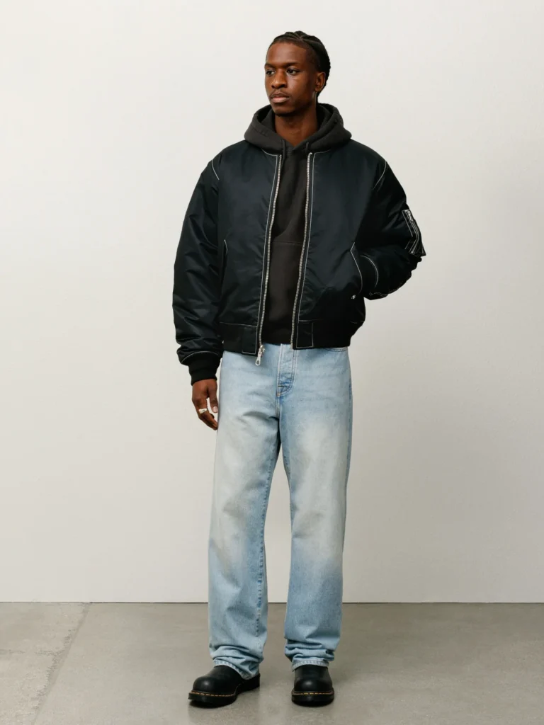 Male model standing indoors wearing a black bomber jacket over a black hoodie, paired with light wash relaxed-fit jeans and black boots.