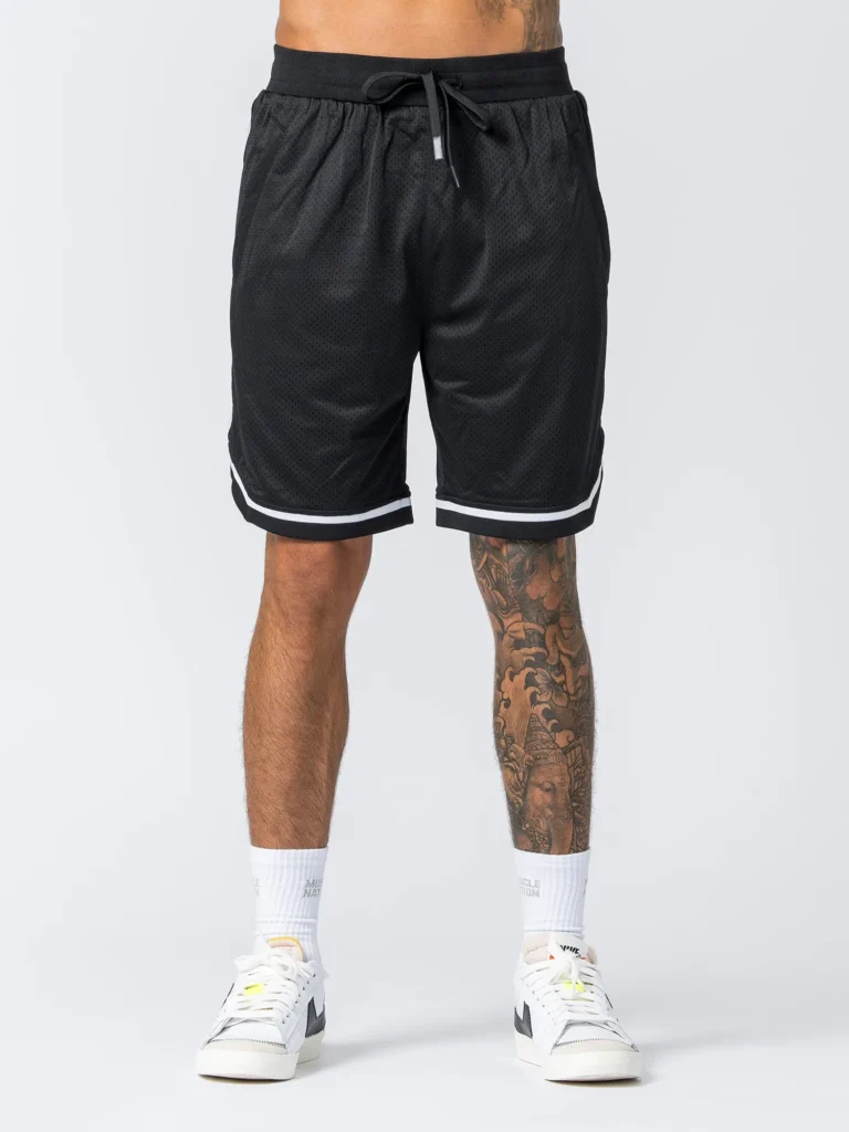 Man wearing black athletic mesh shorts with white trim and a drawstring waistband, paired with white crew socks and white low-top sneakers. A large leg tattoo is visible.