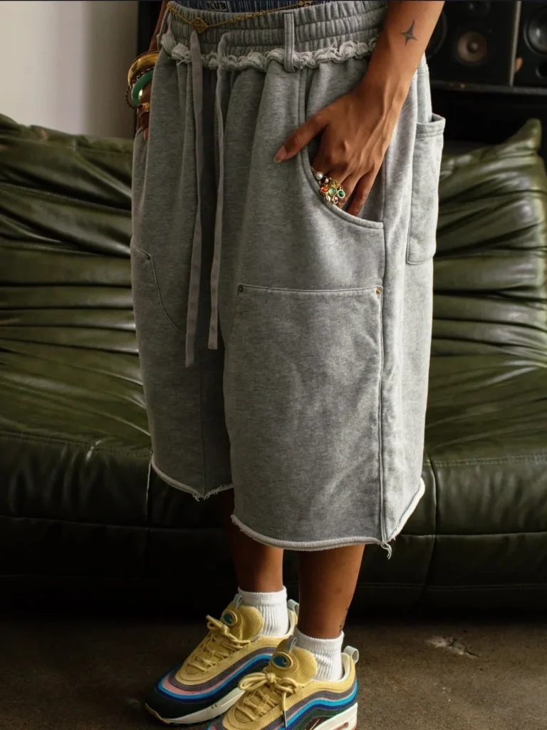 Person wearing baggy gray sweat shorts with raw hems, large front pockets, and drawstring waist, styled with colorful sneakers.