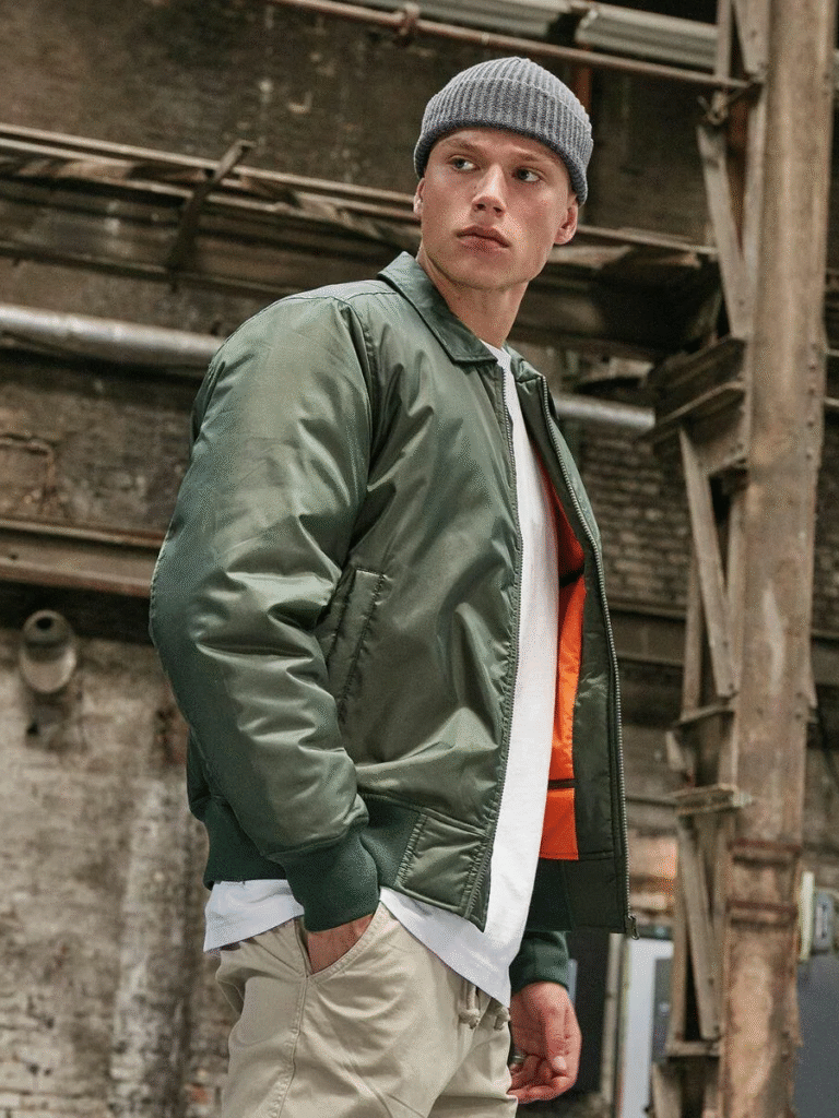 Man in an olive green bomber jacket, white t-shirt, beige pants, and a gray knit beanie, standing in an industrial setting with a confident expression.