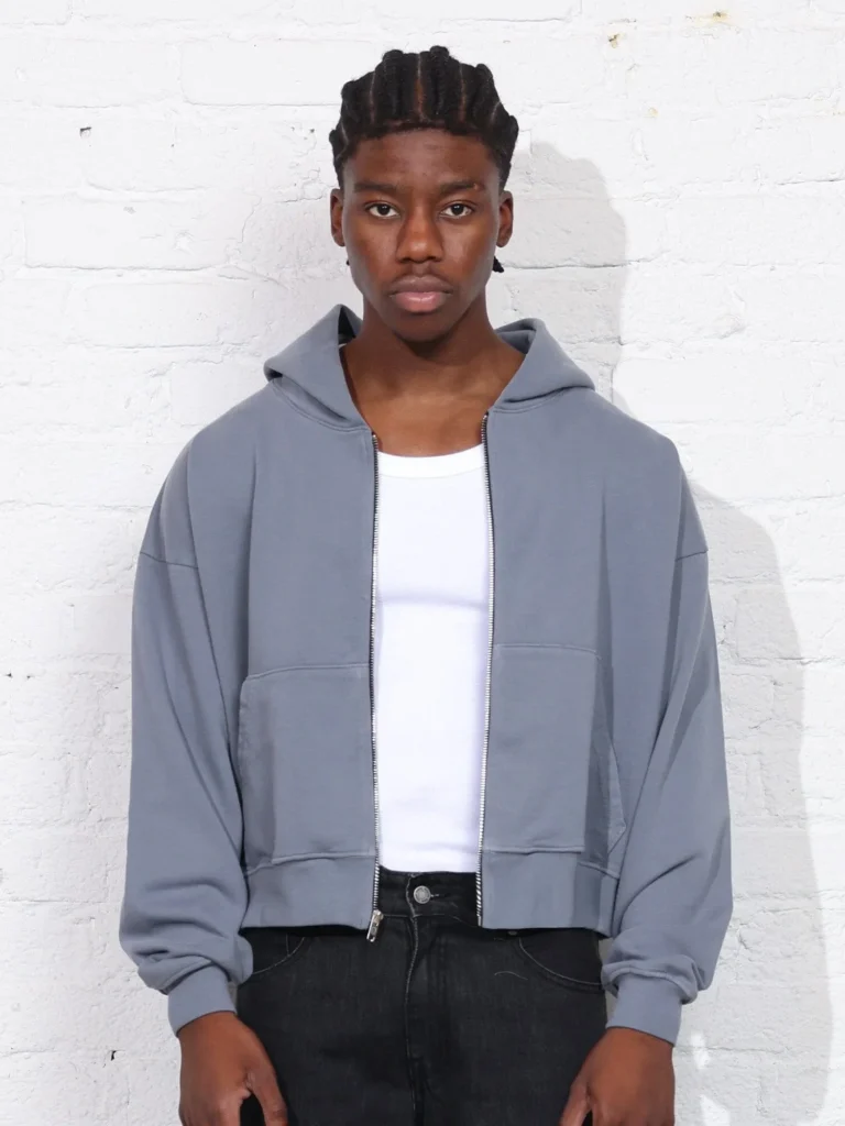 Man wearing light gray cropped zip-up hoodie over white tank top and black jeans against white brick wall