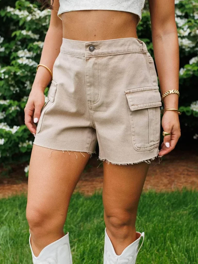 Woman wearing high-waisted beige cargo shorts with raw hems, styled with a white crop top, gold jewelry, and white cowboy boots, standing on grass with greenery in the background.