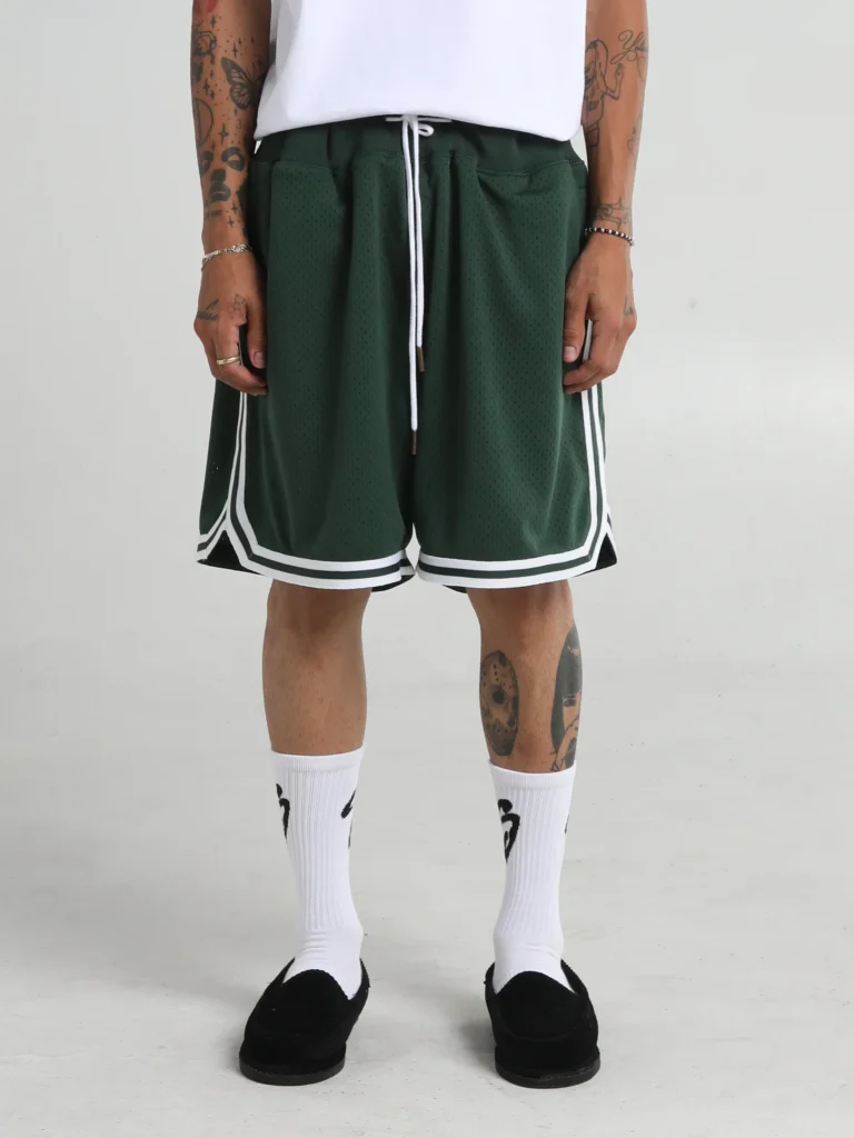 Man wearing oversized dark green mesh basketball shorts with white trim and a long drawstring, paired with a white T-shirt, white crew socks with black graphics, black slip-on shoes, and visible arm and leg tattoos.